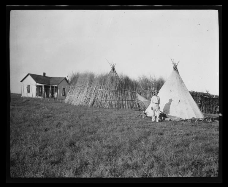 Man in a Yard with Two Tipis, a Thatched Fence, and Small House